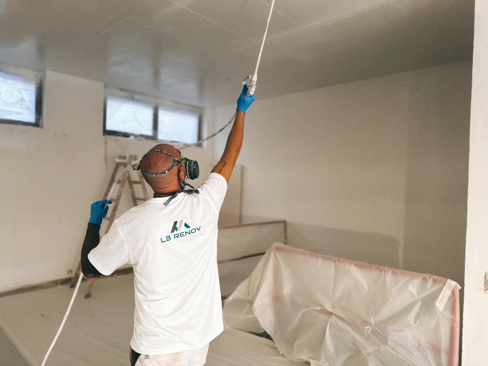 Un peintre de l'équipe LB RENOV pulvérise un plafond, masque et gants, mobilier protégé sous bâche.